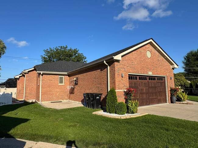 Ground-level view of a newly installed dark gray/black asphalt shingle roof on a red brick home with an attached garage under a clear blue sky. The photo highlights the new roofline, white fascia, and clean gutters. The brown wood-look garage door and well-maintained front lawn emphasize a high-quality exterior roof renovation.
